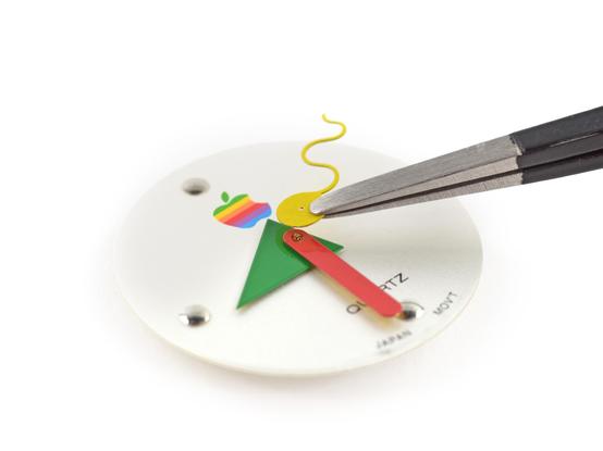 The second hand, which is yellow and squiggly, being held with tweezers above the mechanism that holds all the hands on the Apple analog watch. The minute and hour hands, which are red and green, respectively, are still mounted to the face. The Apple rainbow logo is present above the hands, and the text "Quartz" and "Japan Movt" in all caps below the hands.