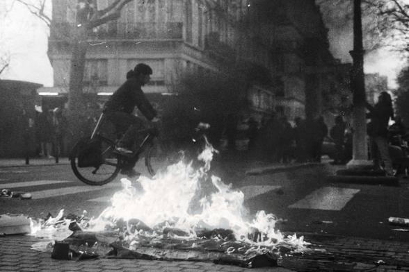 Une scène urbaine en noir et blanc. Au premier plan, des flammes s’élèvent d’un feu sur la chaussée. Un cycliste passe à vélo sans ralentir ni tourner la tête. En arrière-plan, des passants et des silhouettes floues. Atmosphère tendue, contrastée par le calme du cycliste.