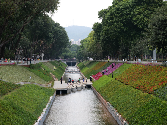 A stream runs straight forward with flowers and grass sloping up on either side. In the middle there is a crossing with a number of people on it 
