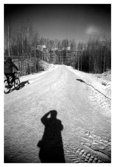A wintery but sunny shot from a snowy walkway. Bare trees and residental buildings are in the background, a brave winter cyclist just entered the scene from the left. The shooter's shadow fills the foreground.