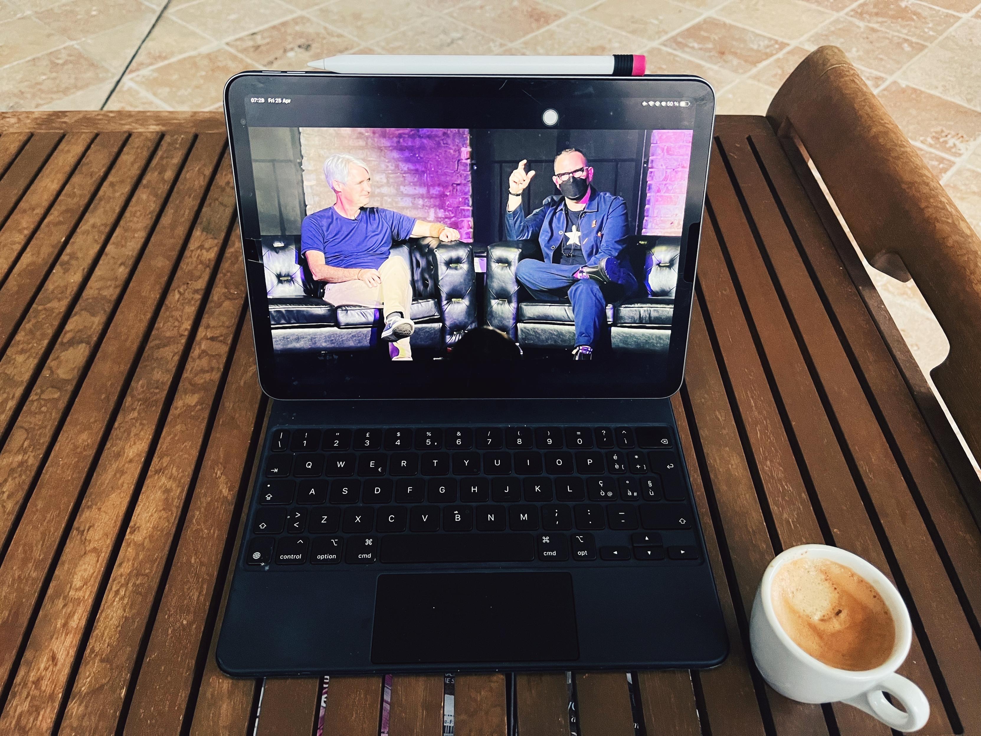 A photo of an iPad sitting on a table showing on screen a frame from Cory Doctorow’s conversation with Mike McCue at Fediverse House - they’re sitting on stage next to each other. Next to the iPad there is a small cup of coffee 