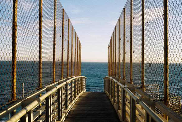 Fotografía de una pasarela vallada al atardecer. Al fondo se ve el mar, dando la sensación de que la pasarela acabe en el agua. 
Photograph of a fenced walkway at sunset. The sea can be seen in the background, giving the impression that the walkway ends in the water.