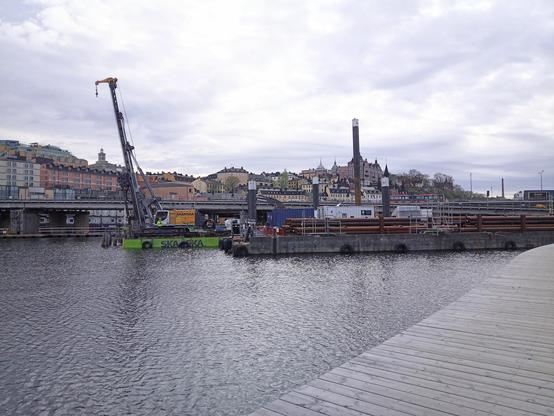 A float with a crane and other reconstruction equipment on a water surface. In the foreground a piece of a wooden walk way, in the background a railway bridge and behind it some buildings on the opposite shore.