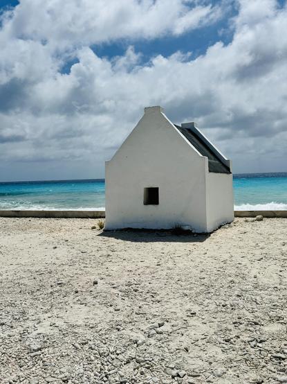 Small, white masonry shelter on a beach. The building looks like a tiny house with white walls and a solitary small square opening, facing the camera. It has a dark, wooden, angled roof like a house. The sand around it is white and the skies are partly cloudy. The sun is shining and the blue water of the ocean is behind it.