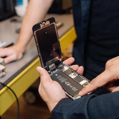 iPhone with its screen flipped upwards at a 90 degree angle, exposing the phone's internals. The phone is being held by someone's left hand, and they're using their right index finger to point at one of the phone's internal components. In front of the person is a workbench, and in the background is another person who appears to be looking at the phone.