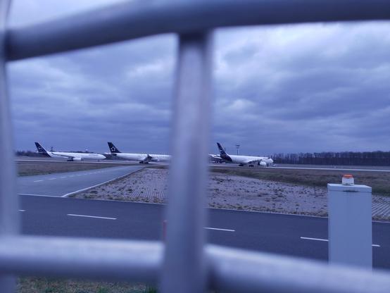 Drei geparkte Flugzeuge der Lufthansa, seitlich durch einen Zaun fotografiert.