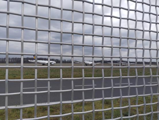 Drei geparkte Flugzeuge der Lufthansa, durch einen Zaun fotografiert.