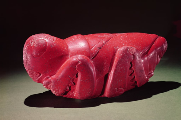 a sculpture,  it is of a grasshopper, very anatomically accurate, carved in stone, it is red colored, against a dark background , it is facing left , seen from the side 