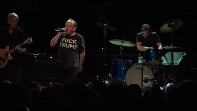 Duane Denison (guitar), David Yow (voice) and Mac McNeilly (drums) on stage. David Yow wears a black T-shirt with the words FUCK TRUMP in white colour