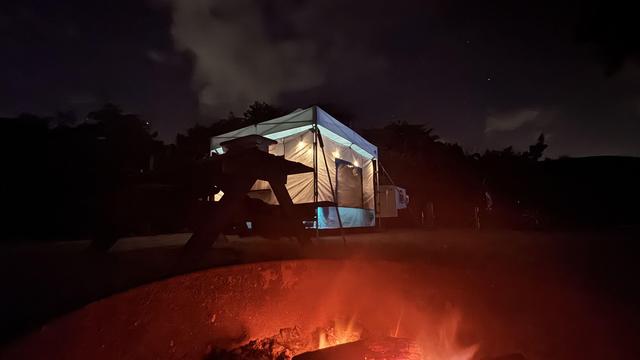 Campfire with a lighted tent and starry night sky in background.