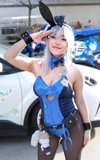 Young woman in cosplay.  She is wearing a blue bunny suit with ears in a police theme.  The suit is two toned blue with a heart shape cutout just below the cleavage line.  The inner material is a light blue from the breast line down through through the crotch area.  A darker blue wraps around the sides and back.  Cleavage is clearly visible as a result of transparent straps.  Around the neck is a light blue collar with a small dark blue bow.  Around the waist is a thin black belt from which is a small black pouch and anime plush figures.