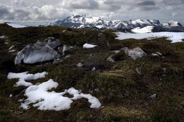 Les Alpes suisses. Au premier plan, des plaques de neiges fondent avec l’arrivée du printemps. Quelques rochers ancrés se dressent, clairs, dans l’herbe rase et sombre d’un début de printemps. En haut de l’images, beaucoup plus loin, les hautes sommets des Alpes tutoient les nuages. 
