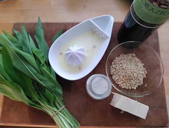 Küchenbrett aus Holz auf dem ein Büschel Bärlauch liegt, dazu ein Stück Parmesan, Glasschüssel mit gehackten Mandeln, einem Hlas Salz, einer Zitronenpresse und eine Flasche Olivenöl.