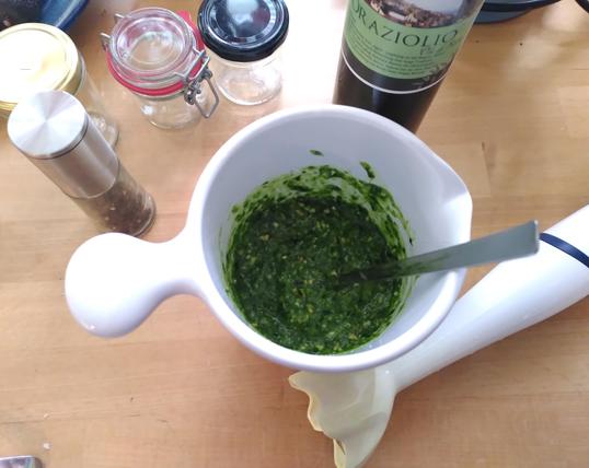 Kunststoff-Topf mit fertigem Pesto, daneben ein Pürierstab, eine Flasche Olivenöl, eine Pfeffermühle und kleine Gläser.