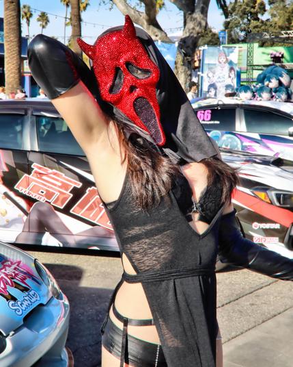 Cosplayer wearing a red ghostface mask from the movie Scream.  The mask is covered in gems which sparkle in the sunlight.  She wore a black bra under a thing dress.  Her arms are covered in long black latex gloves and used a metal chain and red collar.  Her arm is up exposing her side and armpit and the face is tilted toward the right toward the armpit.  In the background there are various automobiles with anime characters painted on them.  The image is taken in natural daylight at Anime Impulse in Pomona California on February 15, 2025.