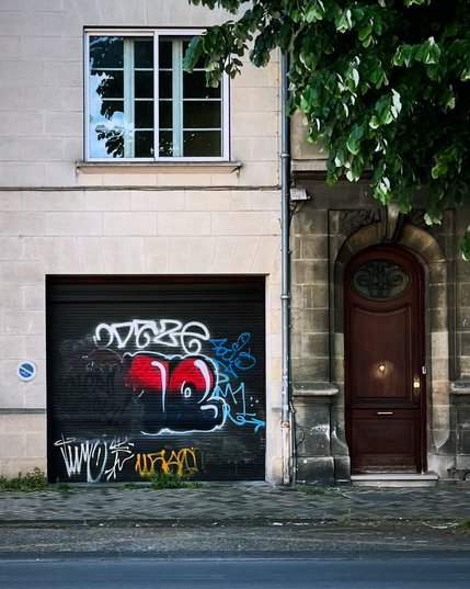 Photo de deux façades de maisons avec le trottoir et la rue partiellement visible. On aperçoit le feuillage printanier d'un arbre en haut à droite. Une porte de garage noire est recouverte de différents tags dont un plus recherché avec le haut des lettres en rouge, le bas en noir, et un effet de relief qui est à moitié effacé, ne laissant que la moitié d'un m et un e.  La composition est rendue plus intéressante par les découpages 2/3 1/3 entre les deux façades et entre les parties de celle de gauche.