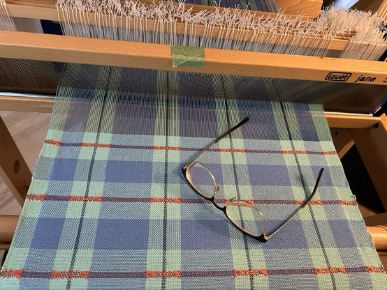 A blue checked teatowel in the process of being woven on a Louet handloom. A pair of glasses sits on the fabric