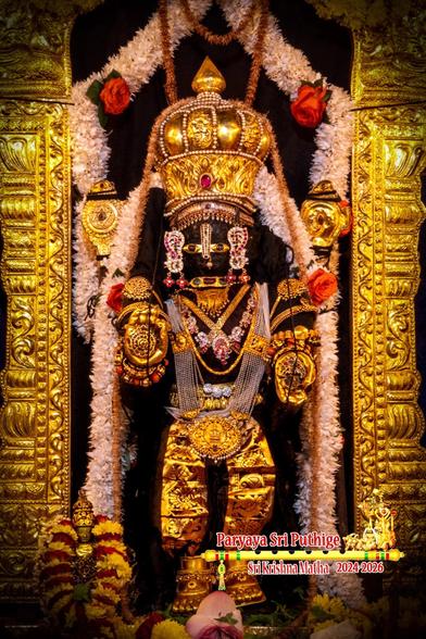 Idol of Hindu deity Krishna with a long draping pearl necklace (haara), gold clothing and ornaments all through (simulating "golden body"), showing a disc in right palm and a crown conch in left palm, wearing a gold crown and shoes, and surrounded by a gold arch. White flower garlands from the top of the arch and around the crown flowing down on either side of Krishna. 