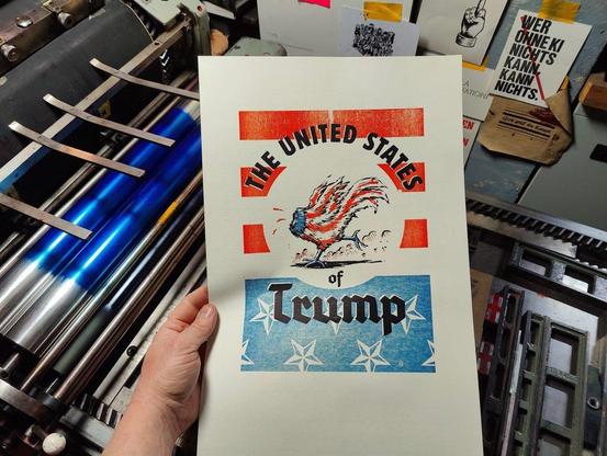 Foto meiner Hand, die ein dreifarbiges Plakat eines kopflos rennenden Gockels in Farben der Vereinigten Staaten von Amerika umrundet vom Schriftzug The United Staates of Trump vor die Druckmaschine hält