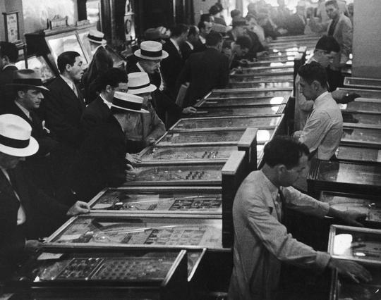 Pinball arcade c. 1935. Several large rows of pinball tables in an amusement parlor are occupied by men, players and spectators alike.

https://www.gettyimages.com/detail/news-photo/men-playing-pinball-machines-in-an-amusement-arcade-usa-news-photo/81599094?adppopup=true