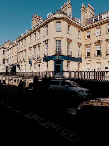 A curved, beige building with multiple windows and a blue door is shown against a clear blue sky. The scene includes parked cars on the street, with a shadowy area in the foreground. The architecture features traditional elements and the neighborhood appears quiet.