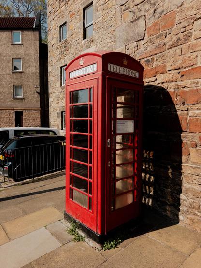 A traditional red telephone booth is situated on a sidewalk next to a stone building. The booth is partially in shadow, and there is a car parked nearby. The setting appears to be a quiet street.