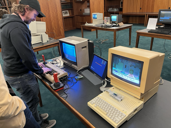 Luke, wearing a hoodie and cap, setting up a Commodore 64 with a raspberry pi with many game titles loaded. A yellowed Apple IIc with color monitor sits in the foreground.