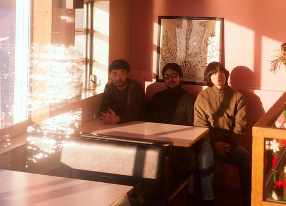 Band photo of the three-piece band Maya Ongaku sitting next to each other at a cosy cafe or diner. The seating arrangement is near a couple of windows allowing natural sunlight to shine inside the space, giving it a warm atmosphere. Photography by Kenta Sawada