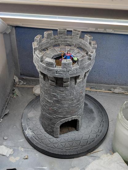 Miniature of a stone tower sitting on a turn table in a spray booth.  There is a hole in the center of the roof and a slipped platform is visible inside it.  The doorway at the bottom shows another slipped platform.  A figure of an elf sorceress wearing blue and purple robes and shooting a greenish fire ball from her hand is standing on the roof facing out over the battlements.