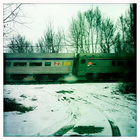 We are stopped at a train crossing, the road is snowy, the image is processed with a white frame and green-cast colour tone.  In front of use, we can see two via train cars, with Canadian flags painted on them, and the cars are slightly skewed to the right because of the motion. 
