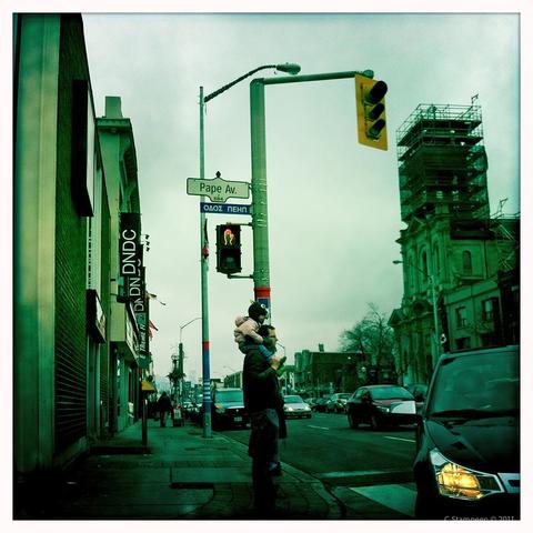 A square image taken with a photo app, white border, aged colour toning.  We are turning a corner of a busy street, the car behind us is very close, and a man is standing, facing right, waiting for a light to turn, with his child sitting up on his shoulders.  The sign post says Pape Street, with the name underneath being written in Greek.  It's a cloudy day.