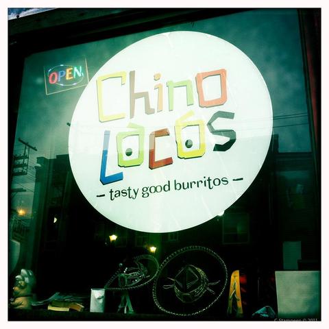 A square image taken with a photo app, with a white border and an aged colour toning.  It's a restaurant store front with a huge white circle sign that says Chino Locos - tasty good burritos - in colourful test.  To the upper left is an open sign, with colourful lights.  Underneath, we can see some dark sombreros with the brims outlined with bright dots. 