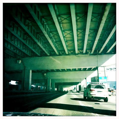 An image taken with a phone app, square, with white border, and aged toning.  We are in the middle of 3 lanes, under an overpass, separated from oncoming traffic by a metal barrier.  Light is coming from the right, creating shadows caused by the overpass and the cars ahead.
