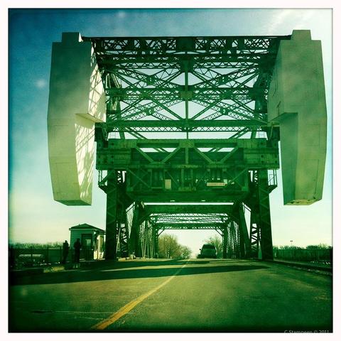 Square image taken with a phone photo app, with a white border and aged colour toning.  We are on a two-lane road, about to go over a bridge.  The bridge has a lot of metal work on top, part of allowing it raised when needed, to allow ships to pass through.