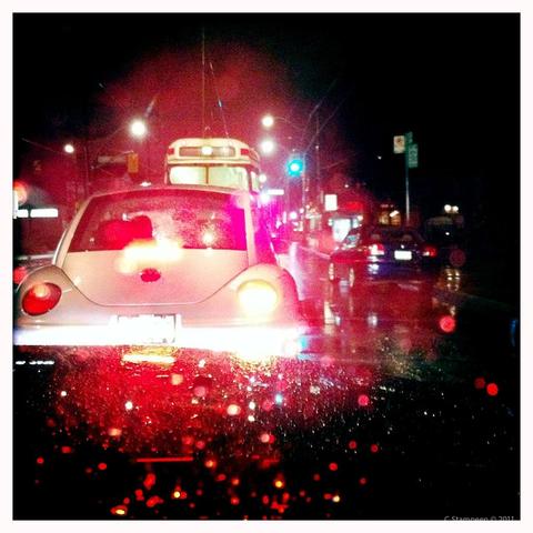 Square image taken with a phone camera app, white border, with a dark vignetting. It's a rainy night, raindrops on the windshield, there is a car in front of us and a street car in from of it.  The traffic light is green, but the image is mostly a red glow. 