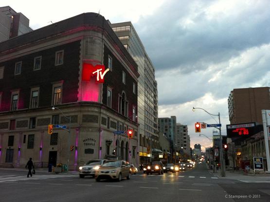 A photo taken as a passenger on a very cloudy day.  We are stopped at a red light, though the blurred vehicles coming towards us suggest some drivers were running the yellow light.  On the left hand corner, a big lit sign is mounted high up on the corner of the building, with a red "M" and smaller white "tv" incorporated in the M. Lower down, the corner also has a sign for Masonic Temple. 