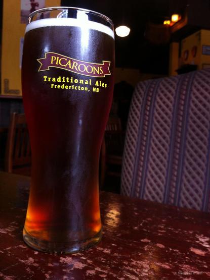 A vertical image of a draught glass of beer.  The beer is dark with a white head.  The glass reads Picaroons, traditional ales, Fredericton, NB. 