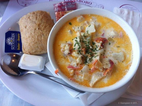 Looking down on a plate that contains a bowl of seafood chowder, with a fresh roll and packaged crackers and butter, sitting on a diner paper placemat.
