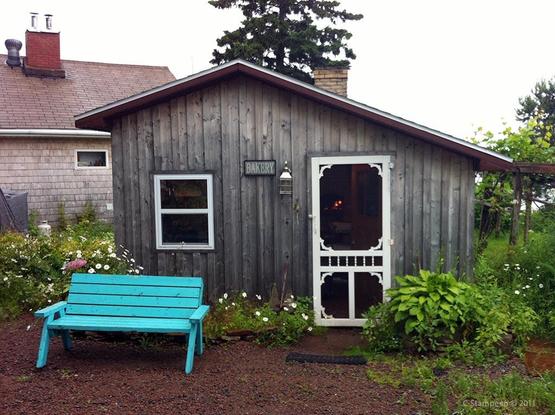 Looking at a cute wooden building with a white screen door to the right and a white framed window to the left.  In front of it, under the window, is a green bench. A sign between the door and window says "bakery."