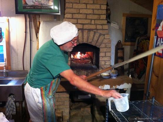 An older man, wearing a green t-shirt, colourful apron and white chef's hat is holding a pizza paddle in his left hand and is putting down a measuring cup with his right.  Behind him is a brick pizza oven and we can see a pizza inside.  