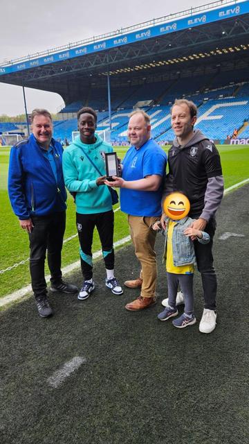 A rectangular glass trophy in a box is handed to Djeidi Gassama by me. There are two other London Owls members and a child stood either side. Everyone is smiling. We're in the manager's box in front of the pitch. The blue seats of the Kop stand are in the background.