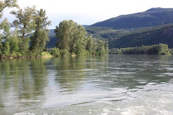 A ripple in the water of a river that flows between rocky and tree lined river banks toward the high hills in the distance.