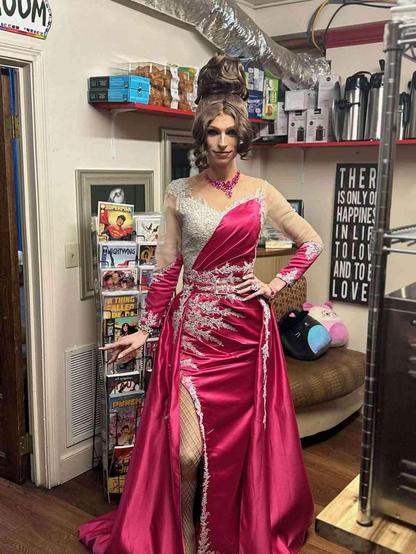 Person in a magenta, embellished, long-sleeved gown, high-fashion hairstyle, and jewelry, standing in a retail space with comic books and food items on shelves.