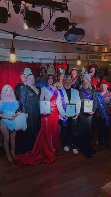 Group of people in elaborate costumes and crowns, holding awards, at a drag pageant.