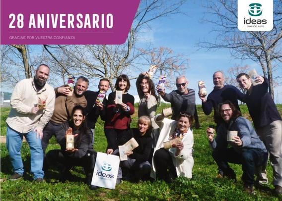 Una foto grupal al aire libre de un grupo diverso de personas sonrientes sosteniendo productos de la marca "Ideas Comercio Justo". El grupo está parado en un campo verde con árboles en el fondo bajo un cielo azul brillante. En la parte superior hay un encabezado que dice "28 Aniversario" y "Gracias por vuestra confianza". El logotipo de Ideas Comercio Justo también está presente en la esquina superior derecha y en una bolsa en el primer plano. Todos parecen felices y celebrando el aniversario de la empresa.