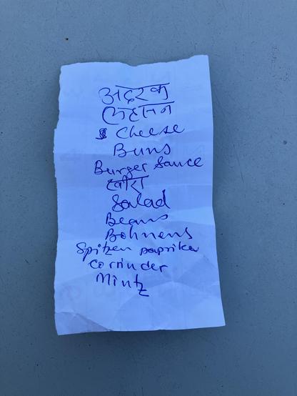 Das Bild zeigt ein zerknittertes Stück Papier mit einer Handgeschriebenen Liste in blauer Tinte auf einem grauen Untergrund. Die Liste enthält verschiedene Artikel, darunter "Cheese Buns", "Burger Sauce", "Salad", "Beans", "Bohnen", "Spitzenpaprika", "Coriander" und "Mint". Oben auf der Liste steht "৩১৬২৫" und darunter "চেস" (Cheese in Bengali). Die Schrift ist unregelmäßig und handgeschrieben, was auf eine persönliche Notiz hinweist.
