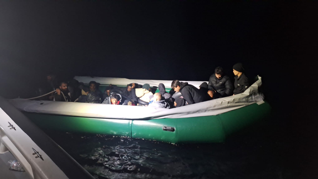 Dunkle Nacht. Von einem Patrouillenboot aus beleuchtet ein Scheinwerfer ein treibendes grünes Schlauchboot, der ober umlaufende weiße Schlauch ist weitgehend luftleer. Im Boot sind knapp 20 Personen zu erkennen; niemand trägt eine Rettungsweste. Eine Person hält eine Leine zu dem Patrouillenboot, von dem aus das Foto aufgenommen wurde.