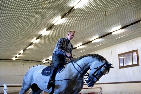 Dans un manège couvert, éclairé au néon, un homme sur un cheval blanc traverse l’image de gauche à droite. 