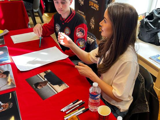 Deux personnes à une table recouverte d'une nappe rouge. L'une des personne tient un stylo au-dessus d'une feuille blanche, l'autre semble discuter avec une autre personne hors champ. Sur la table il y a deux feuilles blanches, une photo de l'actrice dans son rôle ainsi que des stylos ou feutres et une bouteille d'eau.