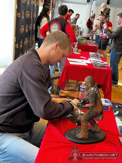 Joonas Suotamo à une table recouverte de rouge. Il signe un document et à sa droite, il y a une statuette de Chewbacca. Dans le fond il y a plusieurs personnes dont des membres de l'équipe de Génération Star Wars.